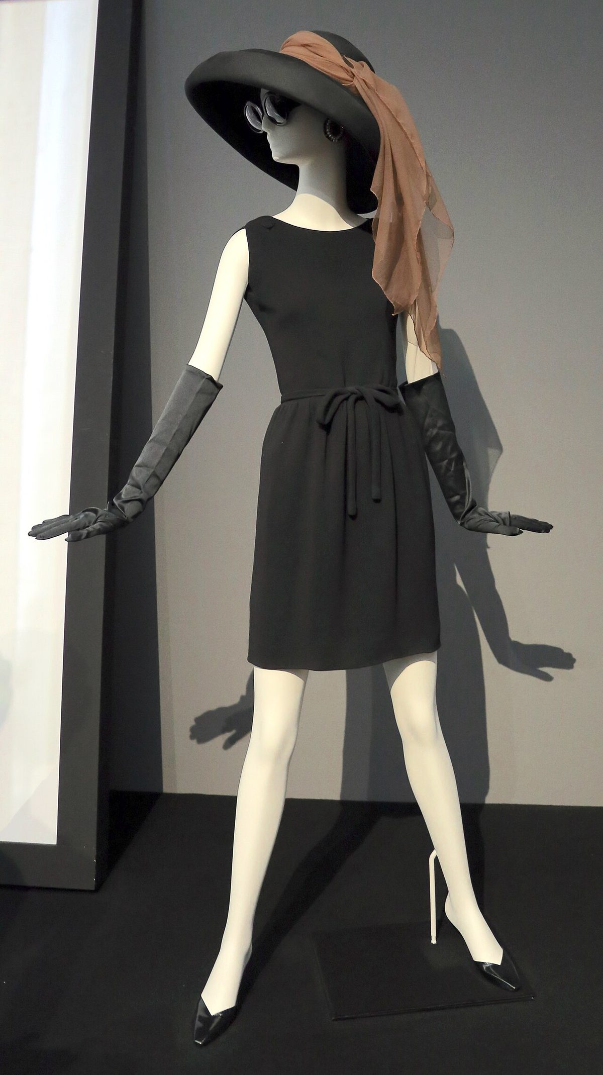 A short black Givenchy dress with a wide-brimmed hat and long gloves, worn by Audrey Hepburn in Breakfast at Tiffany's (1961), shown on a mannequin in a museum display