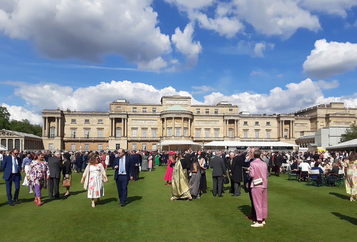 Guests in day dresses, hats and suits on the lawn at a Buckingham Palace garden party, with the palace's garden facade behind them