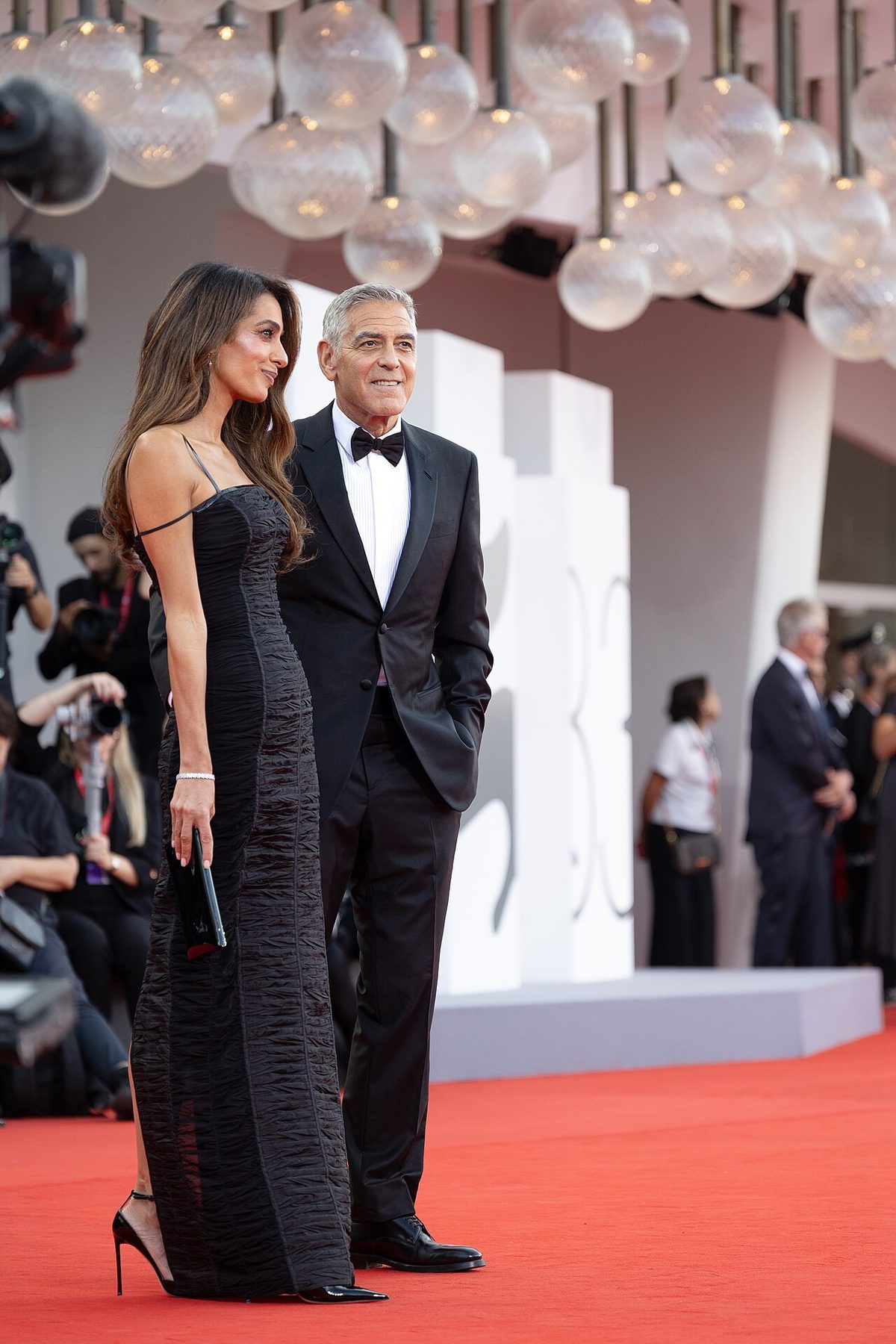 Amal Clooney in a black ruched column gown with George Clooney on the red carpet at the 2026 Venice Film Festival